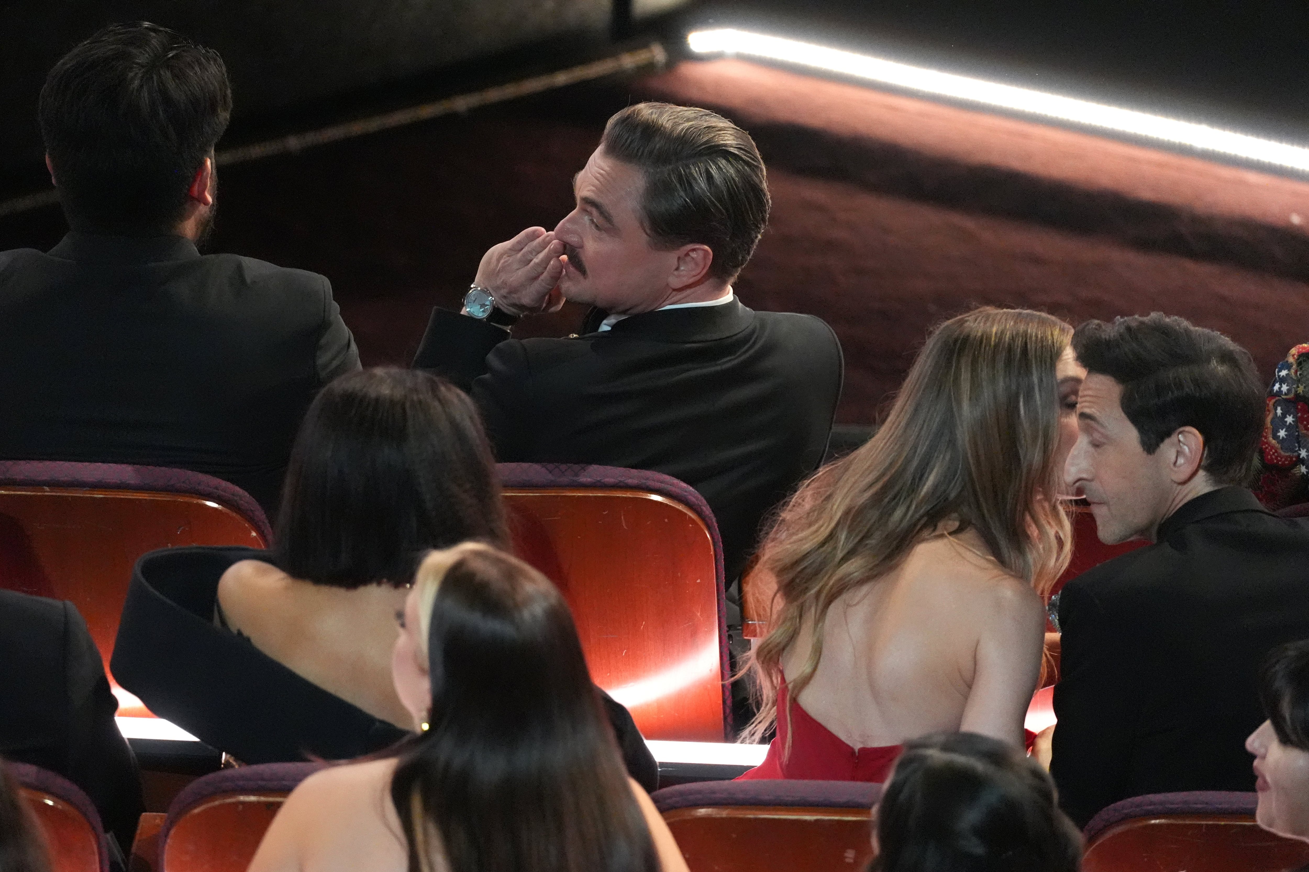 Leonardo DiCaprio mirando la sala llena de figuras mientras la mujer de Adrien Brody, Georgina Chapman, le dice algo al oído a su marido